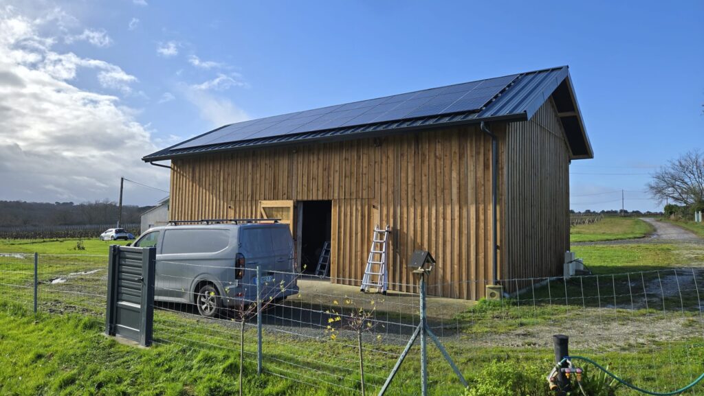 Bâtiment en bois moderne avec bardage vertical et toit métallique intégrant des panneaux solaires photovoltaïques. Van de chantier et échelles dans un vignoble.